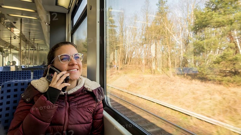 A woman in a cozy jacket is talking on her phone while traveling on a train. She gazes happily out the window at a sunlit landscape, enjoying the scenic journey