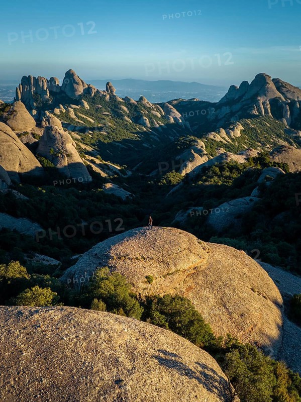 A breathtaking view of Montserrat's rugged rock formations in Catalonia, Spain. The image captures a lone figure standing on a large boulder, surrounded by lush greenery