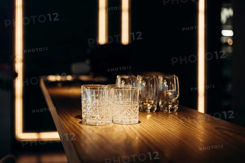 A collection of empty glasses sits on a polished wooden bar, illuminated by warm ambient lighting, evoking a sophisticated and inviting atmosphere