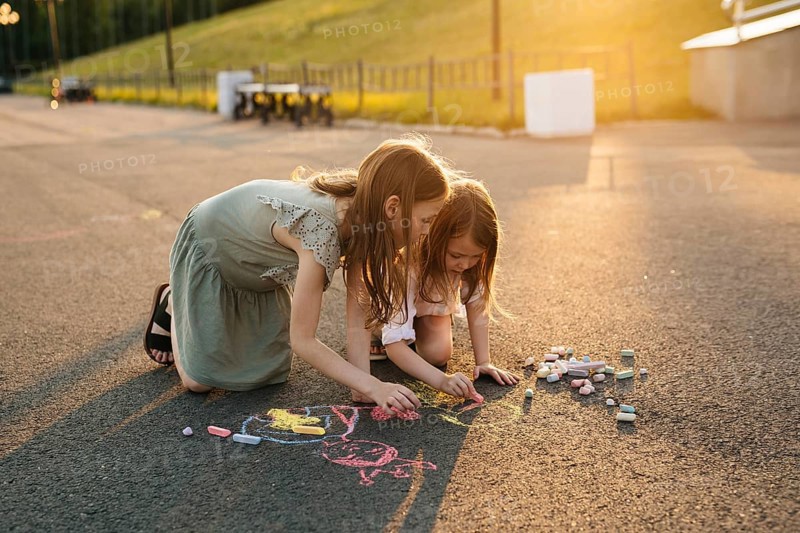 Two kids draw with colorful chalk on asphalt during a bright, sunny day, surrounded by lush greenery. A scene of creativity and childhood joy unfolds in the warm light in summer