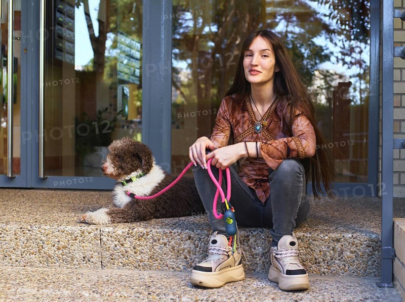 Young woman sitting on steps outside a building, holding a pink leash attached to a fluffy brown and white dog. She is relaxed, wearing casual clothing and sneakers