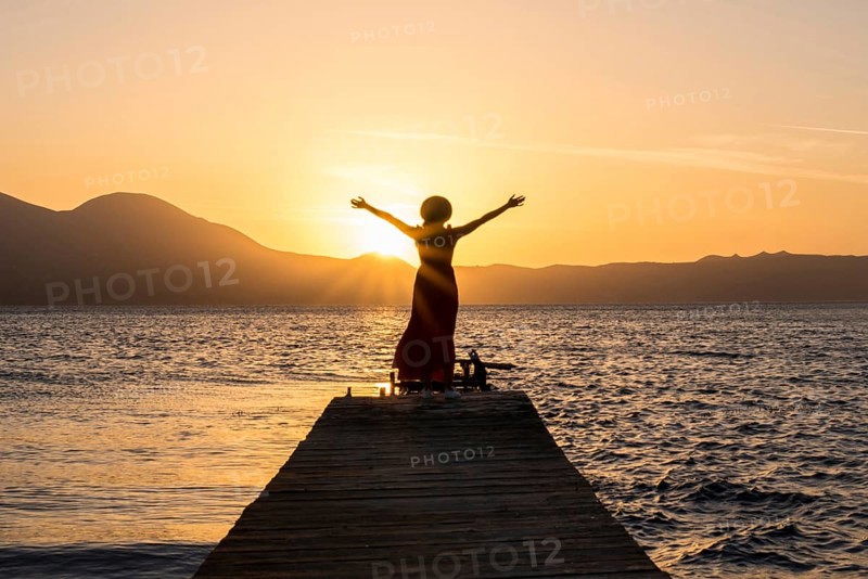 A woman stands on a wooden pier at Papikinou Beach, Milos Island, Greece, embracing the sun as it sets, casting a golden glow over the tranquil sea