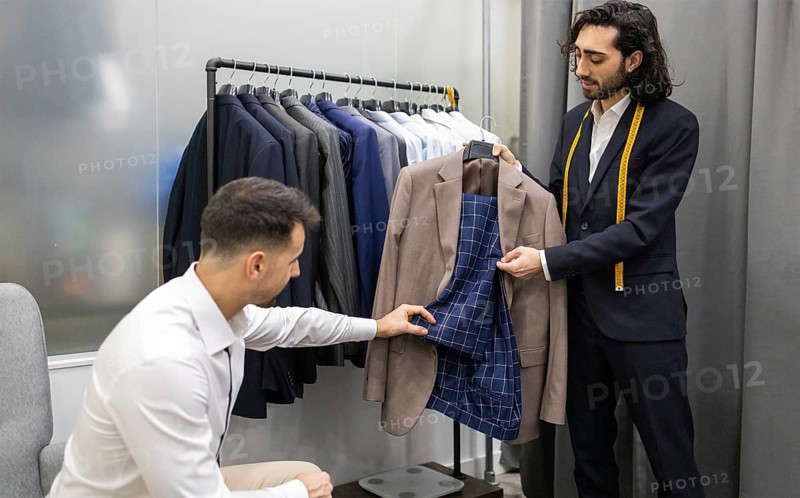 A tailor assists a customer in selecting a suit in modern store. The tailor shows the inner lining of a jacket while the customer examines it. Background features a rack of various suits