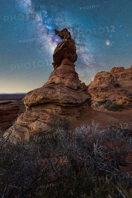 A breathtaking view of a rock formation beneath the Milky Way in Coyote Buttes. The night sky, filled with stars and vivid colors, contrasts beautifully with the rugged, rocky landscape below
