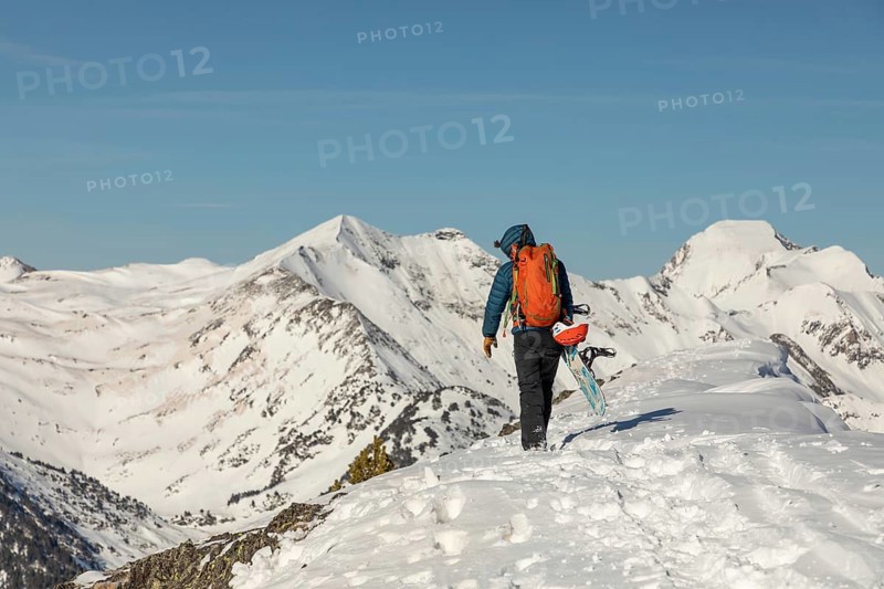 Back view of an unrecognizable person hiking up a snow-covered mountain with a snowboard. The snowboarder, dressed in winter gear, is surrounded by a breathtaking alpine landscape