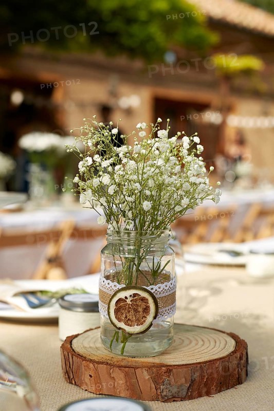 A rustic wedding table centerpiece featuring delicate white flowers in a mason jar adorned with lace, placed on a wooden slab. Ideal for romantic boho-themed weddings