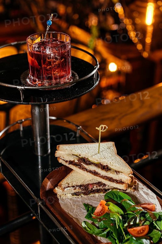 A gourmet club sandwich filled with tender veal cheeks, served alongside a fresh tomato and spinach salad. Accompanied by a classic negroni cocktail for a perfect pairing