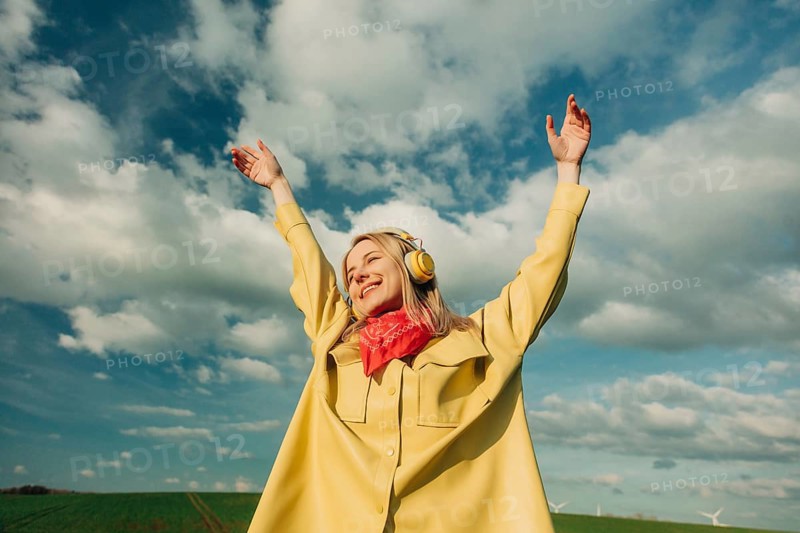 A joyful woman in a yellow coat and red scarf listens to music through headphones, raising her arms under a bright, cloudy sky in an open field, embodying freedom and happiness