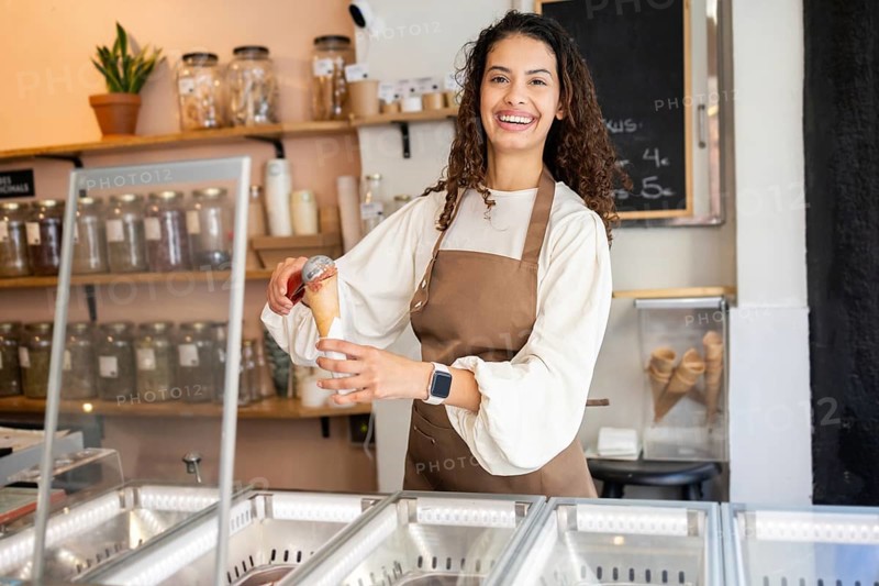 A cheerful woman working in an ice cream shop, serving a scoop of delicious ice cream in a cone. She wears a brown apron and stands behind the counter with joy