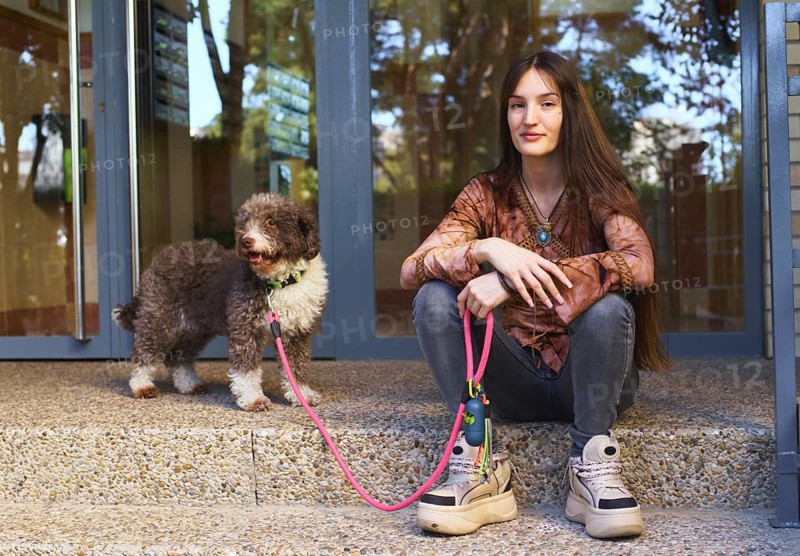 A young woman sits on steps next to a dog with a pink leash, enjoying a sunny day. The scene captures a moment of companionship and relaxation outdoors