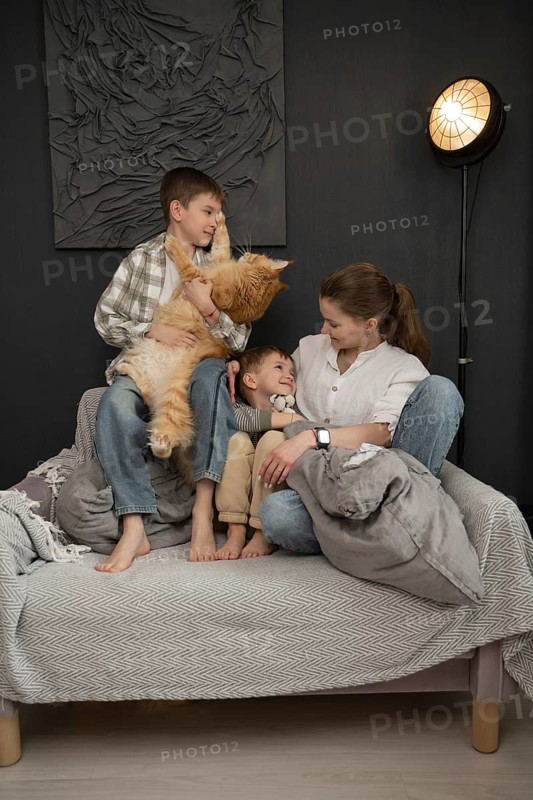 A warm family scene featuring a mother and her two children sharing a joyful moment with their fluffy orange cat. They are seated on a couch in a cozy living room setting