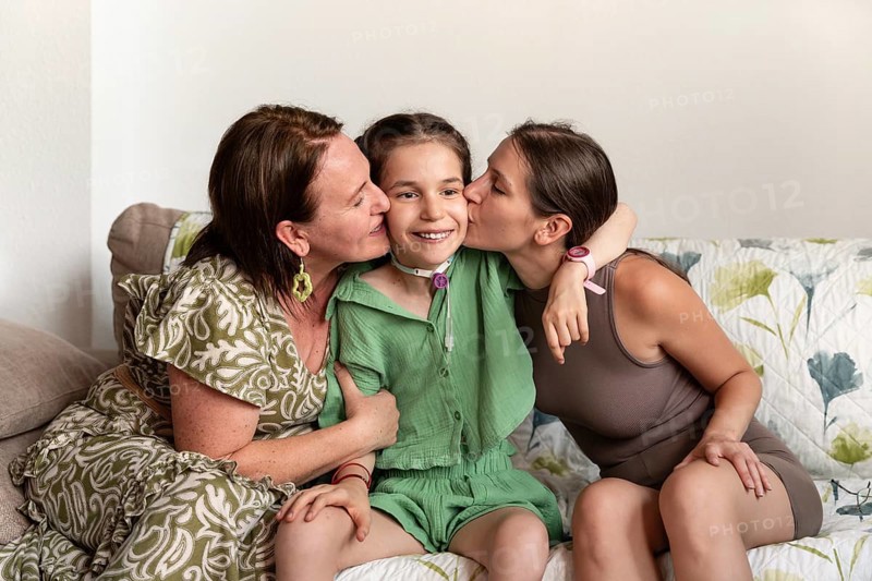 A joyful family moment showing a mother and sister embracing a young girl with a tracheostomy. The emotional connection and support are evident in their smiles and affectionate gestures