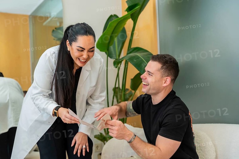 Female doctor in a lab coat smiling and discussing treatment options with a cheerful male patient, reviewing documents during a cordial consultation at an aesthetic clinic