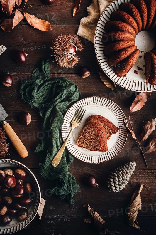 A beautifully styled chestnut cake on a ceramic plate, surrounded by whole chestnuts, leaves, and rustic decor, presenting a warm autumnal setting for a cozy treat