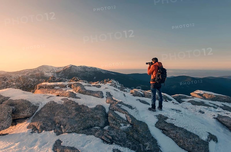 A photographer in an orange jacket captures the snowy landscape of Siete Picos, part of the Guadarrama Mountains, during a picturesque sunset in Madrid, Spain