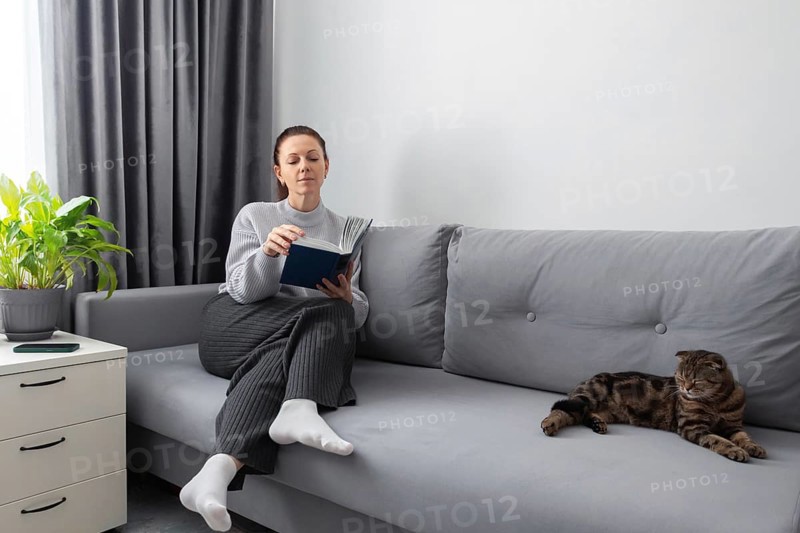 A woman reads a book on a gray couch in a cozy living room with a relaxed cat nearby. The setting features modern decor elements and a potted plant