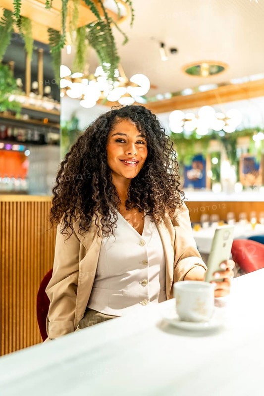 Vertical photo of a happy latin woman using phone while drinking coffee in a modern cafeteria