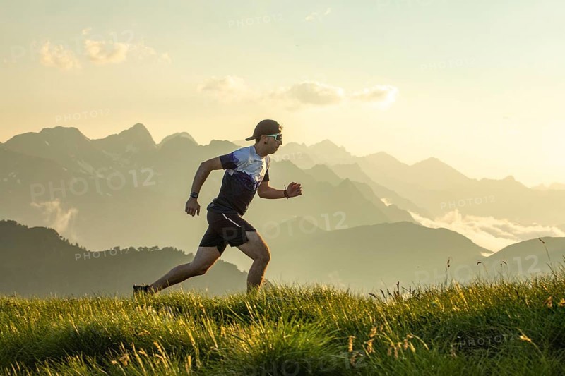Man in athletic gear running on a grass trail with scenic mountain backdrop. The sun sets behind the peaks, casting a warm glow. Wearing a hat and sunglasses, he embodies outdoor activity
