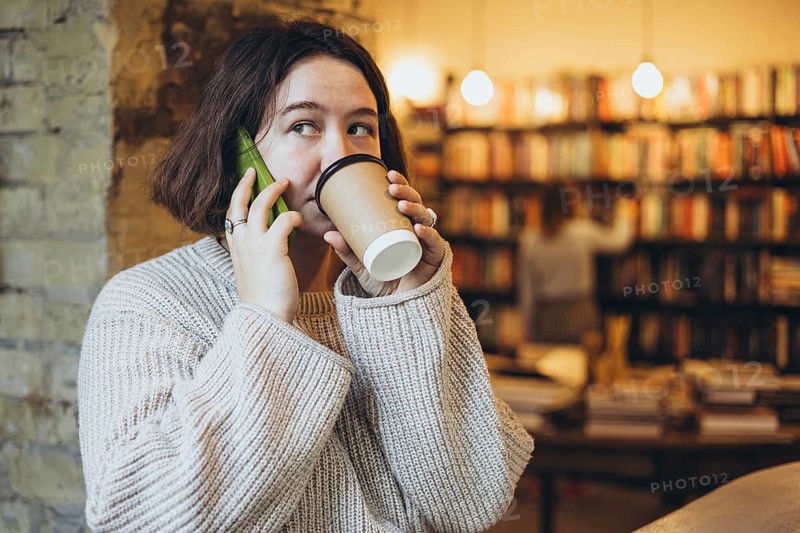 A young woman in a cozy sweater drinks coffee while talking on her smartphone inside a warm bookstore cafe The shelves filled with books create a relaxed and inviting atmosphere