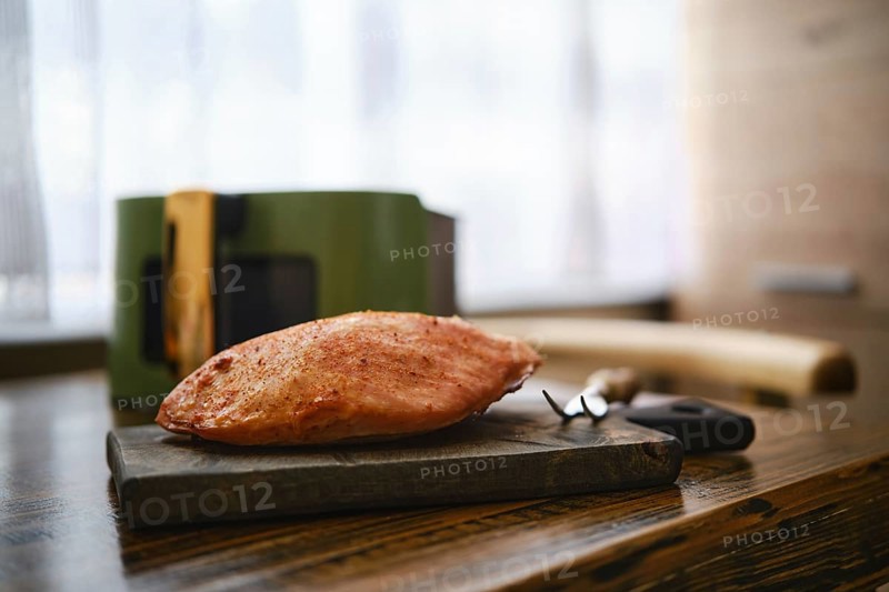 Roasted pork fillet rests on a wooden cutting board in a kitchen table. Air fryer basket and kitchen tools are on blurred background. Natural light fills the room enhancing the cooking scene