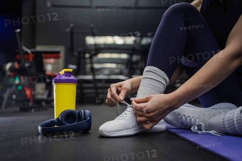A woman ties their white sneakers, preparing for a workout session. Nearby are wireless headphones and a vibrant yellow shaker bottle, ready for gym time