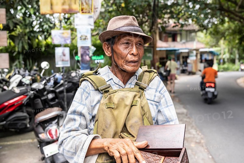 An elderly balinese man wearing a hat and plaid shirt stands on a street, holding a wooden box. Scooters and greenery surround him, creating a vibrant urban scene