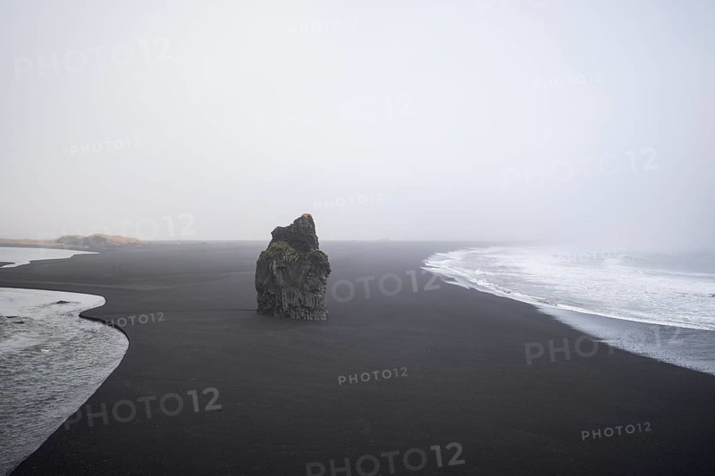 A haunting view of Reynisfjara Beach, Iceland, featuring a solitary rock formation amidst the black volcanic sand under a misty winter sky, captured in solitude