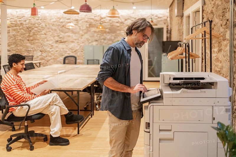 Two professionals in a cozy coworking space, one seated with documents and the other using a copier. The rustic setting exudes creativity and collaboration