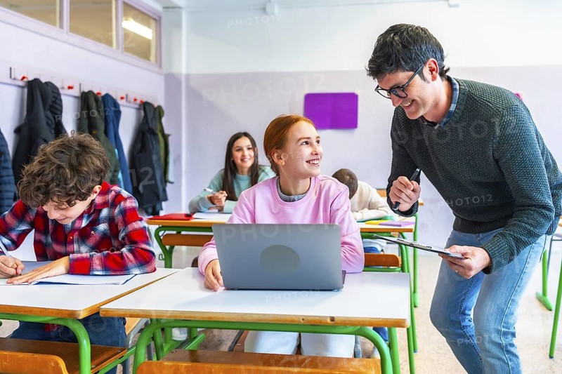 Smiling teacher assisting a student using a laptop in a classroom, other students studying and writing at their desks