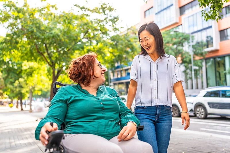Caucasian happy woman with disability using wheelchair talking while walking with an asian friend along the city