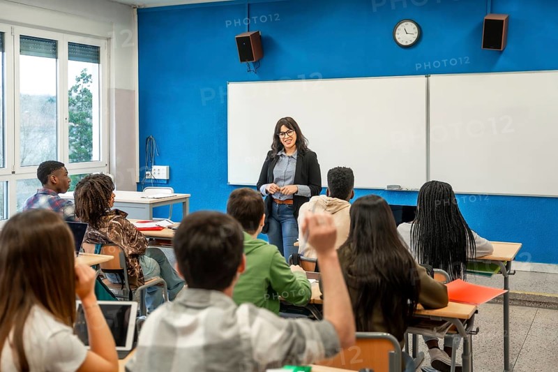 Multiethnic students raising hands during a lesson held by female teacher in high school classroom