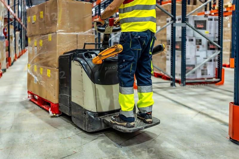 Part of an unrecognizable worker wearing reflective clothes using forklift in a warehouse to move boxes