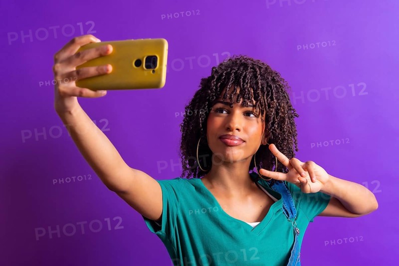 Cheerful young woman with curly hair taking selfie with smartphone and showing victory sign against purple background