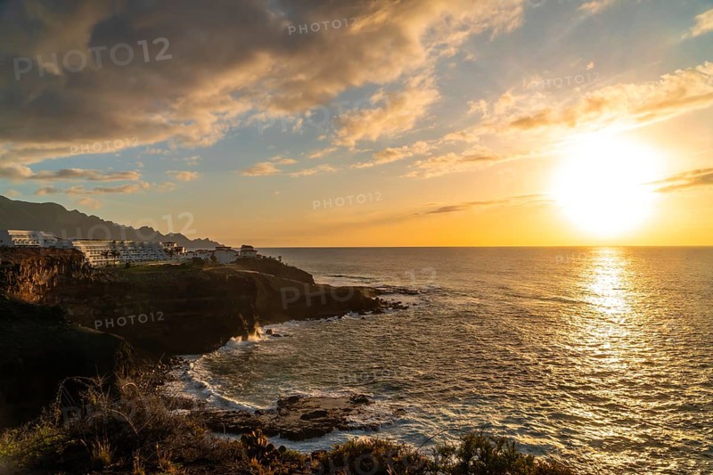 Breathtaking sunset over the atlantic ocean, casting golden light on the rocky coastline and white buildings of agaete, a charming coastal town in gran canaria