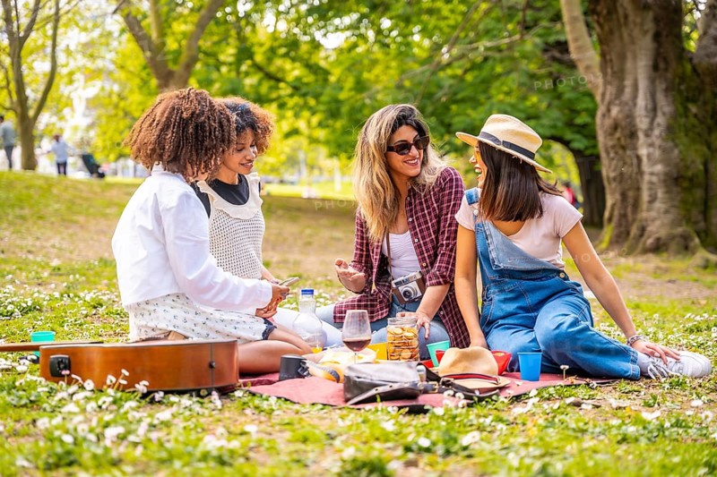 Four female friends having fun, eating and drinking wine at a picnic in a park on a sunny spring day