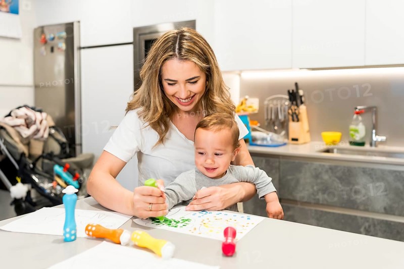 Happy mother and baby boy enjoying creative playtime, painting with colorful dot markers at the kitchen table