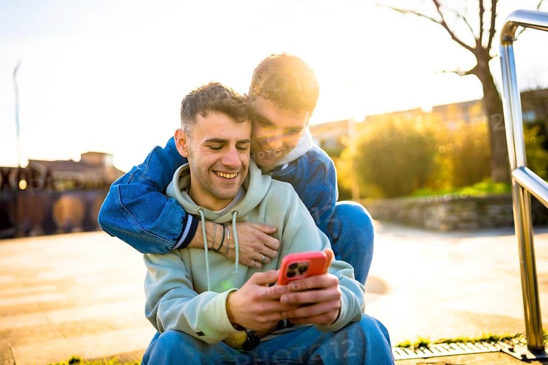 Two men embracing and laughing together while enjoying a smartphone outdoors on a sunny day, radiating joy and affection