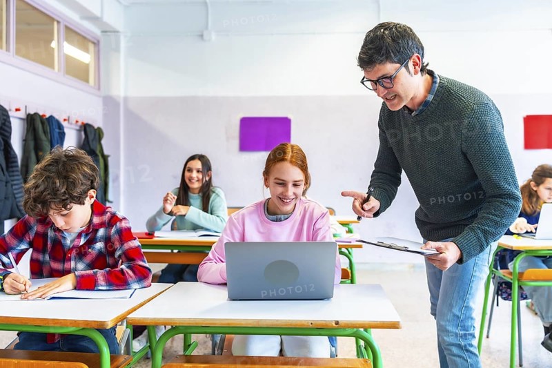 Teacher helping a smiling student using laptop computer in classroom during lesson, education and learning concept