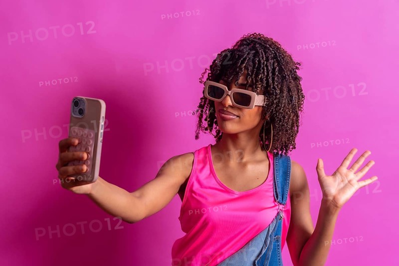 Young woman wearing sunglasses and pink shirt taking selfie with her smartphone while waving with her other hand on a pink background