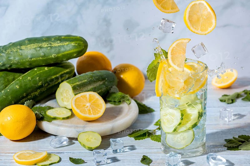 Sparkling cucumber water in a glass with lemon slices, mint leaves and ice cube cubes, with fresh cucumbers and lemons on a table next to it