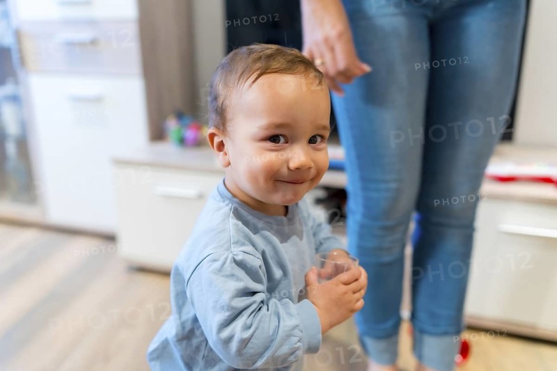 Happy toddler drinking water from a glass, standing close to his mother's legs in a cozy living room filled with warmth and love