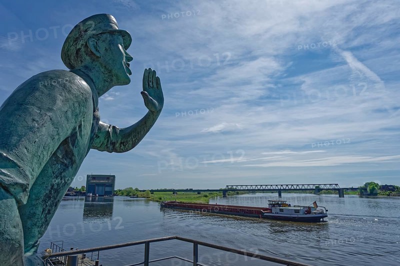 The Lauenburg Caller, a bronze figure by sculptor Karl-Heinz Goedtke, stands in Elbstraße in the old town. The barge Capella is travelling upstream on the Elbe towards the lock. Lauenburg, Schleswig-Holstein, Germany