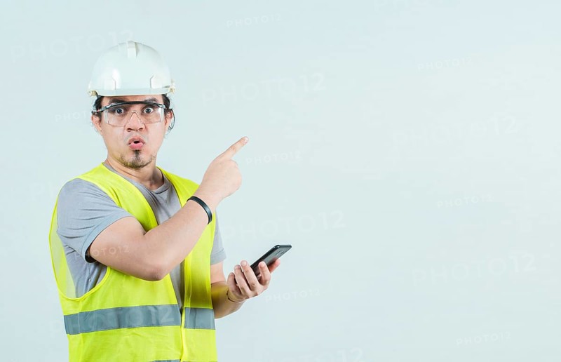 Surprised builder worker with phone pointing at blank space isolated. Amazed engineer holding phone and pointing to the side isolated