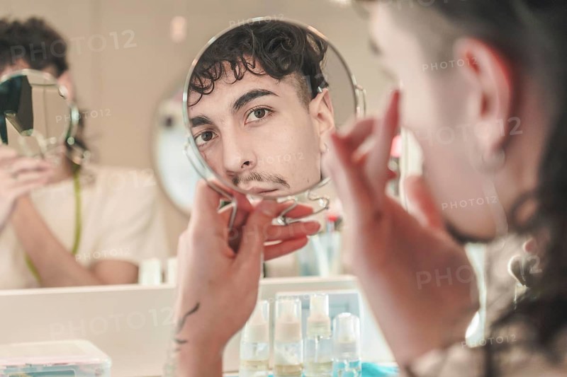Young man checking his face reflected in the mirror while applying creams for his daily skin care routine in the bathroom, with another person out of focus in the background