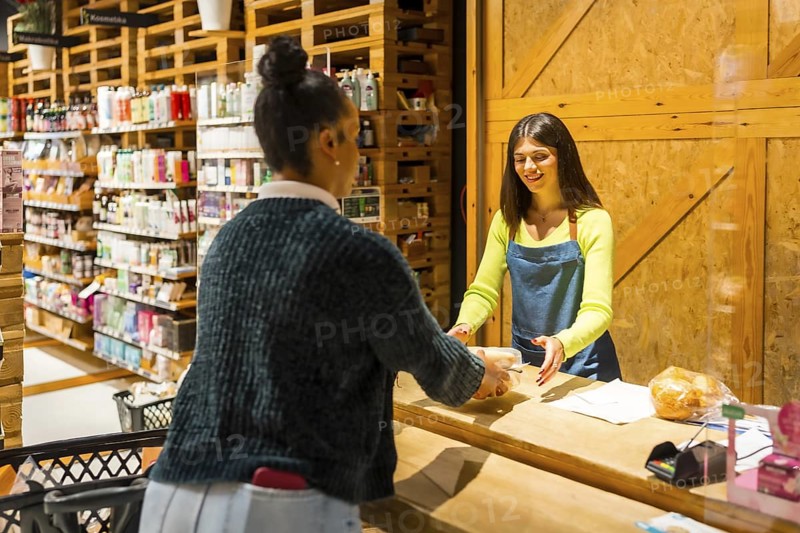 Friendly saleswoman serving a customer buying organic products in a zero waste store or supermarket with wooden furniture