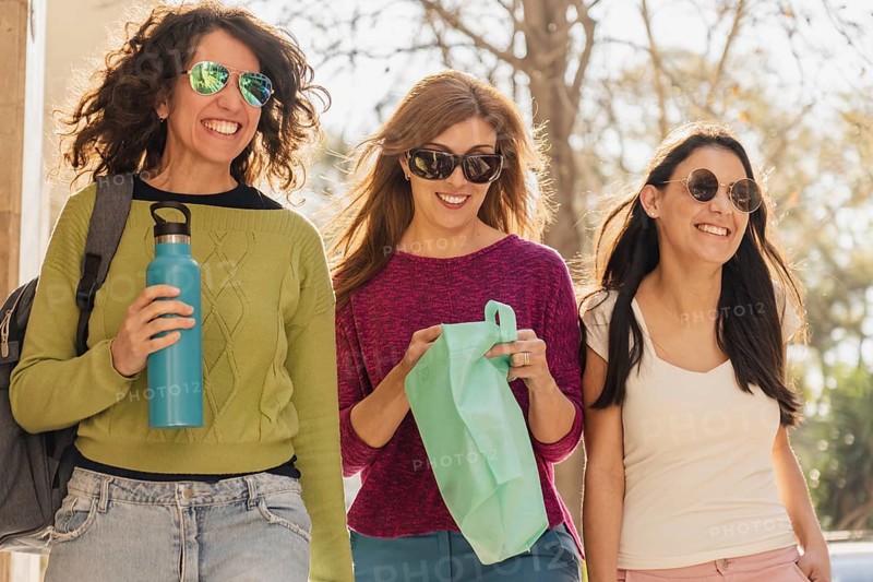 Three women smile and chat while walking outside in the sunlight. They wear casual clothes, sunglasses, and carry eco-friendly bags and a water bottle, enjoying a cheerful and relaxed day