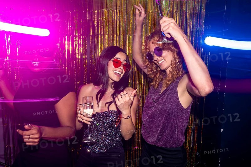 Three cheerful women dancing and drinking sparkling wine at a disco party with neon lights and gold tinsel backdrop