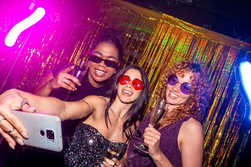 Three multi ethnic female friends are taking a selfie while enjoying sparkling wine at a nightclub, celebrating with neon lights and a gold foil curtain