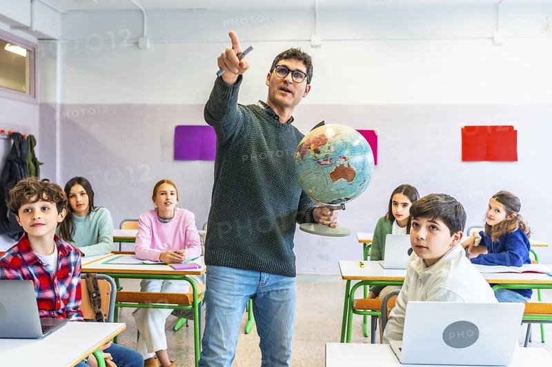 Geography teacher holding globe and pointing at classroom map while teaching geography to attentive students in classroom