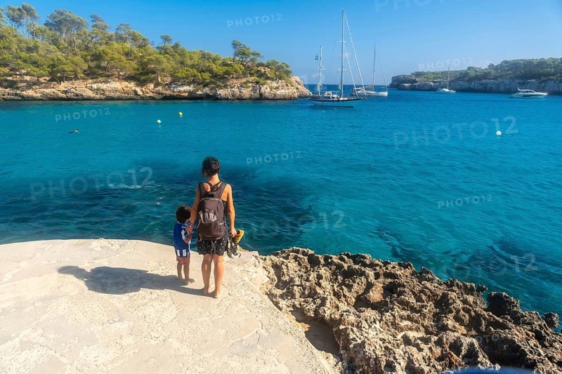 Mother and son standing on a cliff, enjoying breathtaking view of sailboats anchored in the turquoise waters of cala s'amarador, mallorca, spain